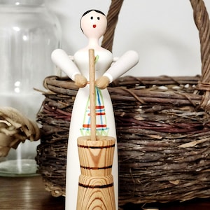 May include: A wooden figurine of a woman in a white dress, holding a butter churn. The churn is made of wood with dark brown bands. The woman has a black painted hairstyle and red lips. A woven basket is in the background.