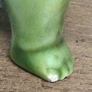 May include: A close-up view of a single green foot or paw, featuring a smooth, rounded shape with a distinct, light-colored chip or worn area at the very tip of the toe or edge, showing a solid, matte-textured surface throughout the structure.