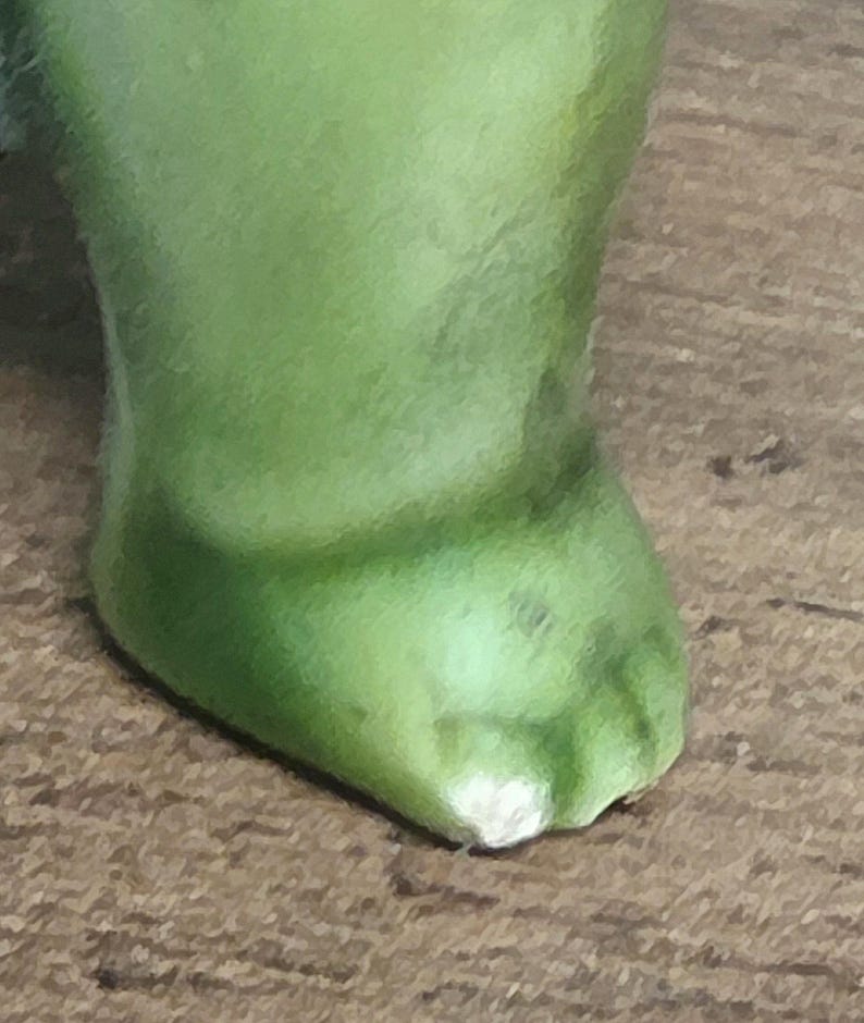 May include: A close-up view of a single green foot or paw, featuring a smooth, rounded shape with a distinct, light-colored chip or worn area at the very tip of the toe or edge, showing a solid, matte-textured surface throughout the structure.
