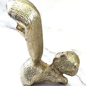 May include: A gold-colored squirrel bottle opener. The opener is in a standing position with its tail curved upwards. The squirrel has a textured surface and a small opening for removing bottle caps. The design is detailed and decorative.