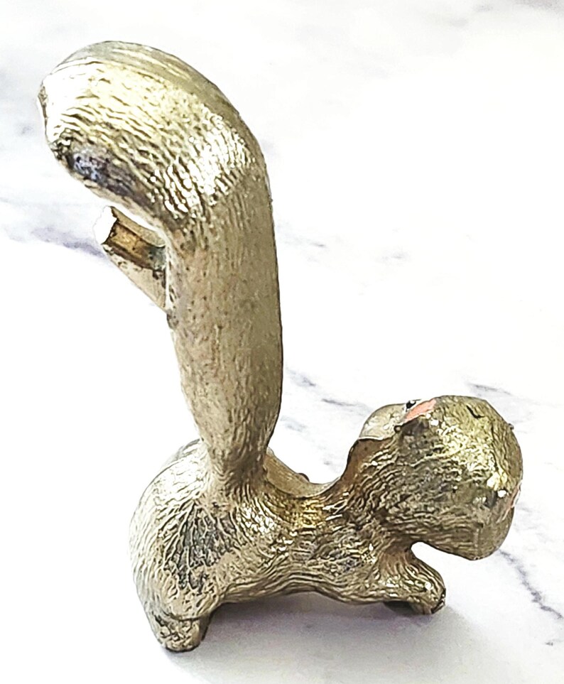 May include: A gold-colored squirrel bottle opener. The opener is in a standing position with its tail curved upwards. The squirrel has a textured surface and a small opening for removing bottle caps. The design is detailed and decorative.