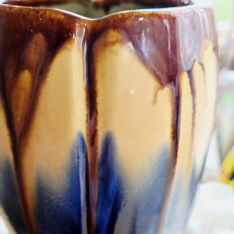 Pode incluir: Um vaso de cer&acirc;mica com um design &uacute;nico. O vaso apresenta uma combina&ccedil;&atilde;o de cores, incluindo castanho, bege e azul. A cobertura castanha escorre pelas laterais, criando um efeito texturizado. O vaso tem uma borda ondulada.