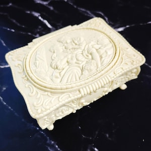 May include: Ivory-colored rectangular jewelry box with an oval relief of two figures. The box has ornate detailing and small feet. The lid features a raised oval with a classical relief scene. The box is made of a solid, opaque material.