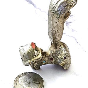 May include: A silver-toned squirrel figurine with a large, curved tail. The squirrel has colorful gemstone accents and a small red accent on its ear. A US quarter is next to the figurine.