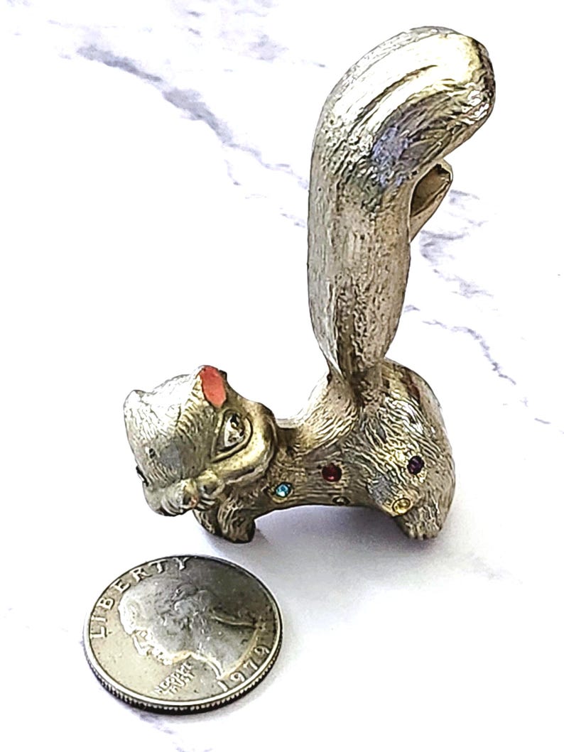 May include: A silver-toned squirrel figurine with a large, curved tail. The squirrel has colorful gemstone accents and a small red accent on its ear. A US quarter is next to the figurine.