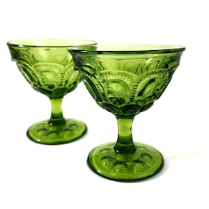 May include: Two green vintage cocktail glasses with a detailed star and crescent moon pattern. The glasses have a stemmed base and a wide, shallow bowl. The glasses are made of green glass and are set against a white background.