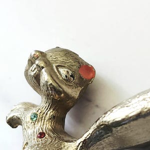 May include: Vintage silver-toned metal brooch in the shape of a stylized animal. The brooch features a textured surface, with red and green jewel accents. The animal has a whimsical design with a large eye and a red jewel on its head.
