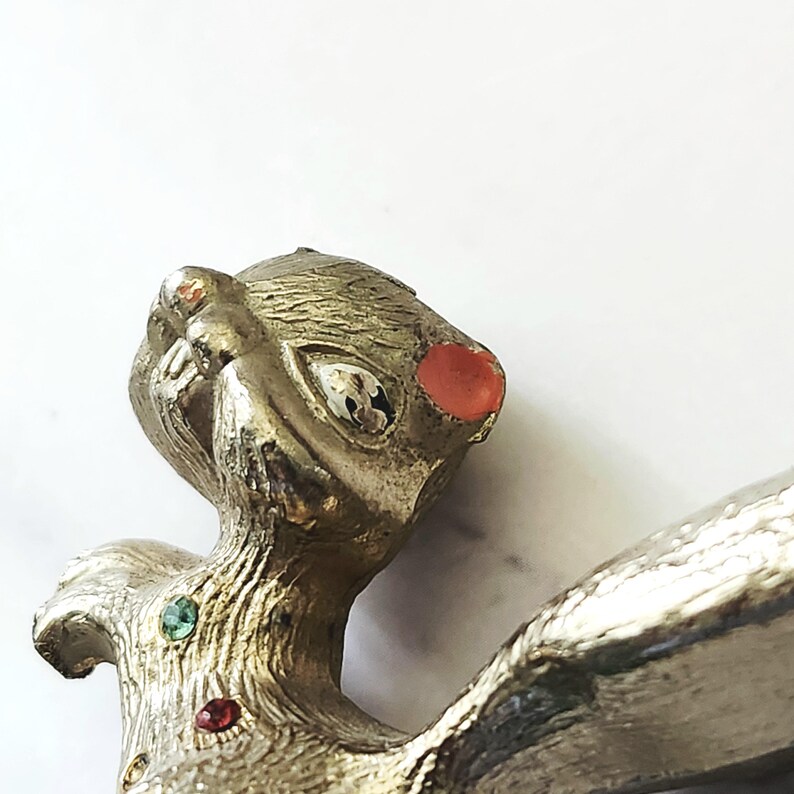 May include: Vintage silver-toned metal brooch in the shape of a stylized animal. The brooch features a textured surface, with red and green jewel accents. The animal has a whimsical design with a large eye and a red jewel on its head.