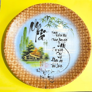May include: Round, woven bamboo tray with a painted scene. The scene features a small house, trees, and a body of water against a light blue background. Calligraphy is written around the scene. The tray has a natural, light brown color.