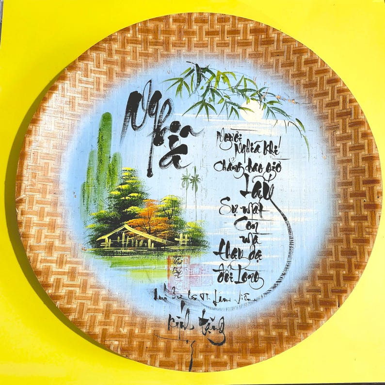 May include: Round, woven bamboo tray with a painted scene. The scene features a small house, trees, and a body of water against a light blue background. Calligraphy is written around the scene. The tray has a natural, light brown color.