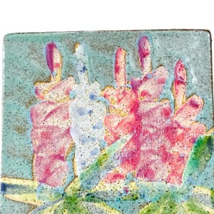 May include: Square ceramic tile with a hand-painted floral design. The flowers are in shades of pink, white, and purple, with green leaves and a light blue background. The tile has a speckled texture.
