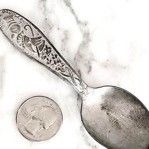May include: Vintage silver spoon with ornate handle details and the inscription "DADDLES". A US quarter coin is next to the spoon for size comparison. The spoon has a worn, antique appearance.