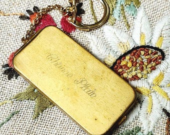 May include: A rectangular, gold-toned change plate with a keychain attachment. The plate has the words "Change Plate" engraved on the surface. The keychain has a chain and a round ring. The background is a floral embroidered fabric.