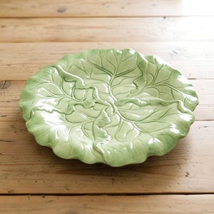 May include: A round ceramic plate featuring a molded design that mimics the appearance of a green cabbage leaf. The surface displays intricate, raised veins and ruffled edges in varying shades of light and dark green with a glossy finish.