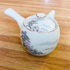 May include: A white ceramic side-handled teapot featuring a hand-painted landscape scene with trees, a mountain, and a small boat. The teapot has a matching lid with a gold-trimmed edge and a long, slender handle extending from the side.