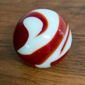 May include: A spherical glass marble featuring a smooth, opaque white base with swirling, bold red bands that wrap around the surface. The object has a glossy, reflective finish and a uniform, rounded shape with no visible seams or markings.