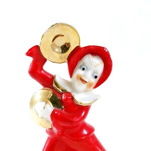 May include: A porcelain figurine of a jester wearing a red and white costume, holding a gold cymbal in one hand and a smaller gold cymbal in the other.