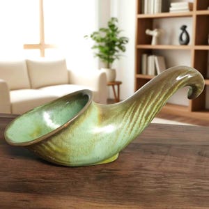 May include: A ceramic decorative bowl in a horn shape, with a light green interior and a brown exterior. The bowl has a curved handle and a textured surface. The bowl is sitting on a wooden table.