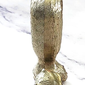 May include: A gold-colored, decorative bottle stopper in the shape of a dog. The dog has a textured surface and a red painted mouth. The stopper is designed to fit into a bottle.