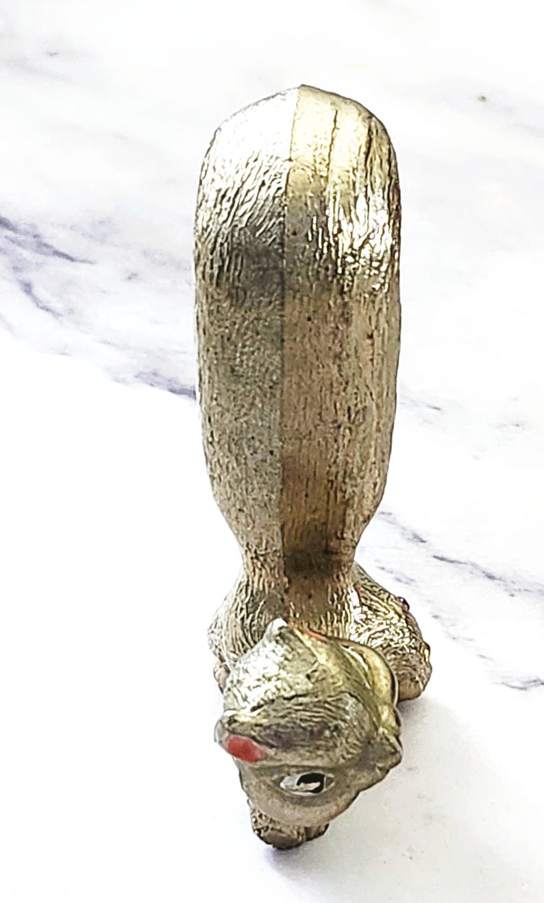 May include: A gold-colored, decorative bottle stopper in the shape of a dog. The dog has a textured surface and a red painted mouth. The stopper is designed to fit into a bottle.