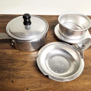 May include: A set of three silver-colored aluminum kitchen items including a small lidded pot with a black knob handle, a deep circular cup-like vessel, and a shallow round dish with a flat handle extending from the side of the rim.