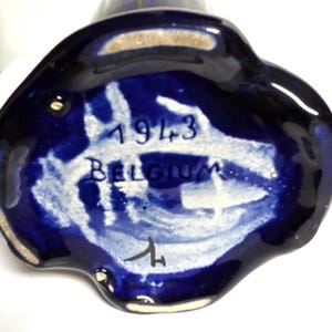 Pode incluir: Um close-up da base de um vaso de cer&acirc;mica azul escuro. A base tem uma forma irregular com pinceladas brancas e azuis. O texto "1943 BELGIUM" &eacute; vis&iacute;vel em branco. Uma assinatura tamb&eacute;m est&aacute; presente.