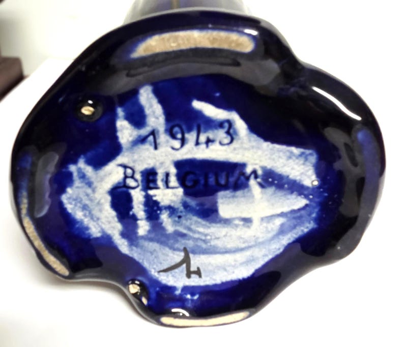 Pode incluir: Um close-up da base de um vaso de cer&acirc;mica azul escuro. A base tem uma forma irregular com pinceladas brancas e azuis. O texto "1943 BELGIUM" &eacute; vis&iacute;vel em branco. Uma assinatura tamb&eacute;m est&aacute; presente.