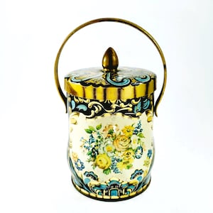May include: Vintage metal container with a lid and handle. The container is cylindrical with a cream-colored background and floral design in yellow, blue, and white. The lid and handle are gold-colored.
