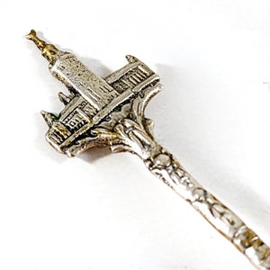 May include: A silver-toned souvenir spoon featuring a detailed architectural design at the spoon's head. The spoon's handle has an ornate, textured pattern. The top of the architectural design has a gold-colored accent.