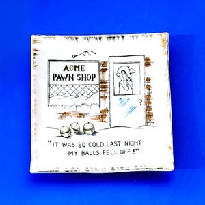 May include: Square ceramic dish with a humorous cartoon scene. The dish features a pawn shop sign, a figure in a doorway, and three round objects. The text reads, "IT WAS SO COLD LAST NIGHT MY BALLS FELL OFF!"