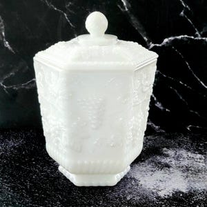 May include: White milk glass candy jar with a lid. The jar has a hexagonal shape and is decorated with a floral design.