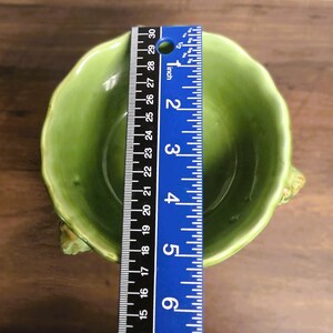 May include: A light green ceramic bowl with a scalloped rim and textured sides, positioned beneath a blue ruler that shows a diameter of approximately 5.5 inches. The bowl has a smooth, glossy interior finish and a rounded, organic shape.