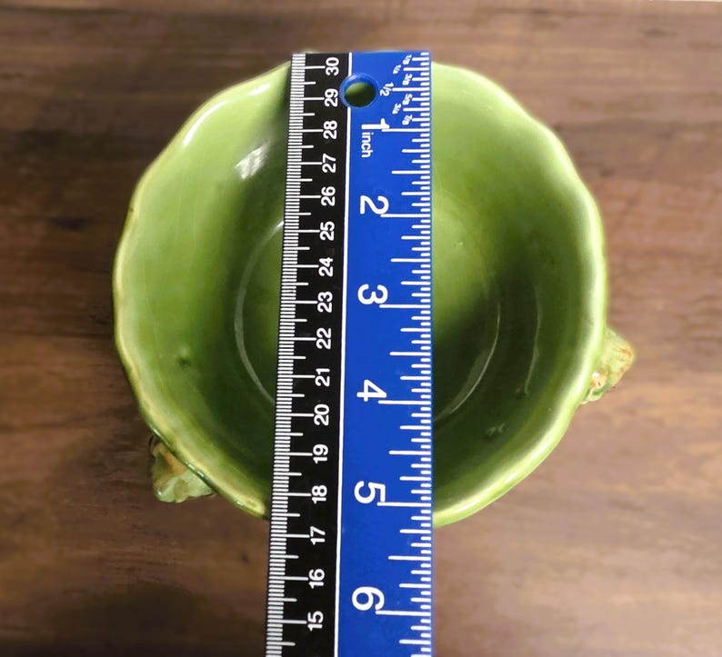May include: A light green ceramic bowl with a scalloped rim and textured sides, positioned beneath a blue ruler that shows a diameter of approximately 5.5 inches. The bowl has a smooth, glossy interior finish and a rounded, organic shape.