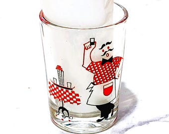 Pode incluir: Um copo de shot de vidro transparente com uma ilustração gráfica de uma pessoa com bigode vestindo uma camisa de bolinhas vermelhas e brancas e um avental branco, ao lado de uma mesa com uma toalha de mesa xadrez vermelha e branca.