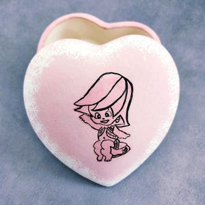 May include: A heart-shaped trinket box with a pink exterior and a white rim. The box features a black and white cartoon illustration of a small figure. The interior of the box is a light beige color.