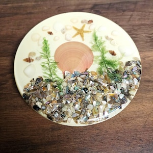 May include: A round, cream-colored coaster with a beach theme. It features a large, pink seashell, a yellow starfish, small seashells, green seaweed, and colorful pebbles. The coaster is made of resin.