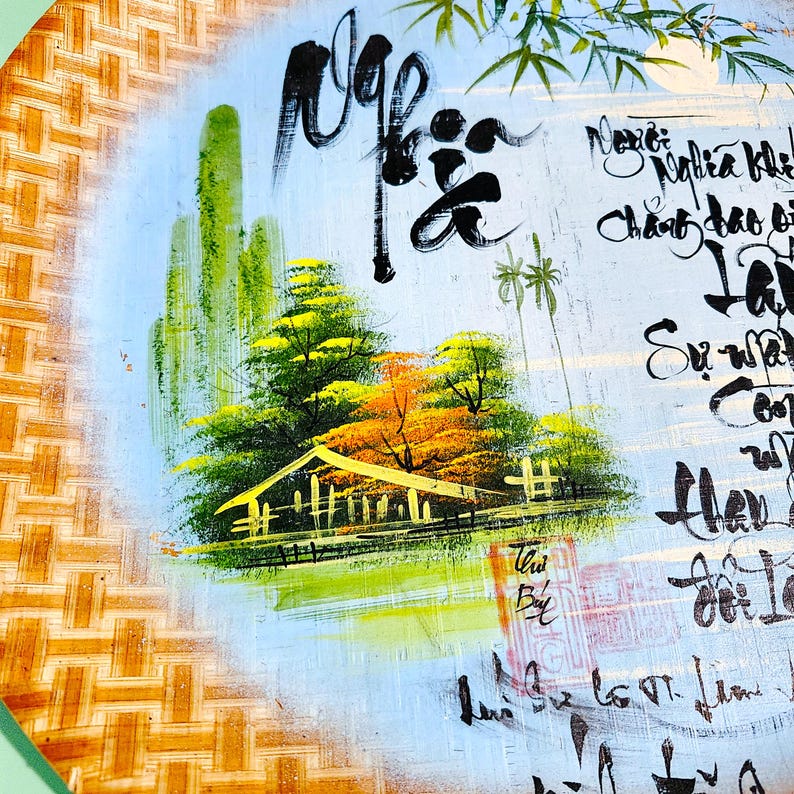 May include: A round, hand-painted artwork featuring a landscape scene with a house, trees, and calligraphy. The artwork is set against a light blue background with a woven bamboo border. The painting uses green, yellow, orange, and black colors.