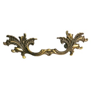 May include: A brass-toned metal drawer pull featuring an ornate, symmetrical design with scrolling leaf-like patterns and a central shell-shaped motif. The hardware has a weathered, antique finish with dark highlights in the recessed areas.