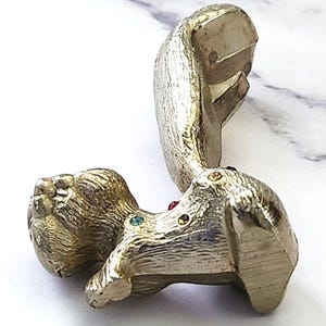 May include: A gold-toned metal squirrel figurine with a textured finish. The squirrel has a long tail and is adorned with colorful gemstones. The figurine is lying on a white surface.