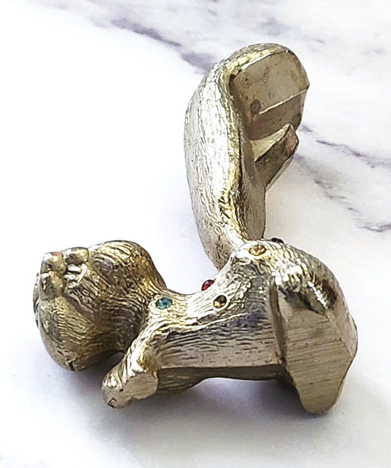 May include: A gold-toned metal squirrel figurine with a textured finish. The squirrel has a long tail and is adorned with colorful gemstones. The figurine is lying on a white surface.