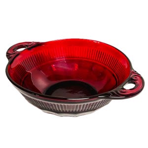May include: A ruby red glass bowl with two ornate handles. The bowl has a ribbed design on the exterior and a smooth interior. The handles have a decorative, swirling design. This vintage bowl is perfect for serving soups or desserts.