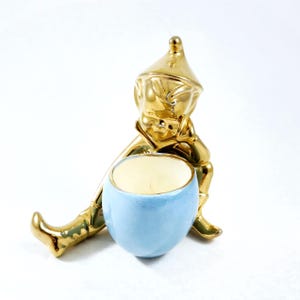 May include: A gold-colored figurine of a seated elf-like character with a blue bowl. The figurine has a shiny, reflective surface. The bowl is light blue with a cream-colored interior. The elf is wearing a hat and boots.