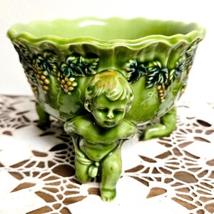 May include: A glossy light green ceramic bowl featuring a scalloped rim and a relief pattern of grape clusters and leaves. The bowl is supported by three sculpted cherub figures that serve as feet, each finished in the same vibrant green glaze.