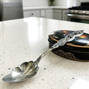 May include: A silver-toned metal souvenir spoon with a shell-shaped bowl and a handle featuring the word NEW YORK in bold letters. The spoon rests against a black oval tin decorated with a colorful floral pattern and gold filigree accents.