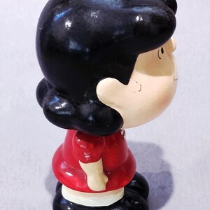 May include: A ceramic figurine of a cartoon character with black hair, a red dress, and black shoes. The figurine is painted with a glossy finish and has a detailed facial expression. The character is looking to the side.