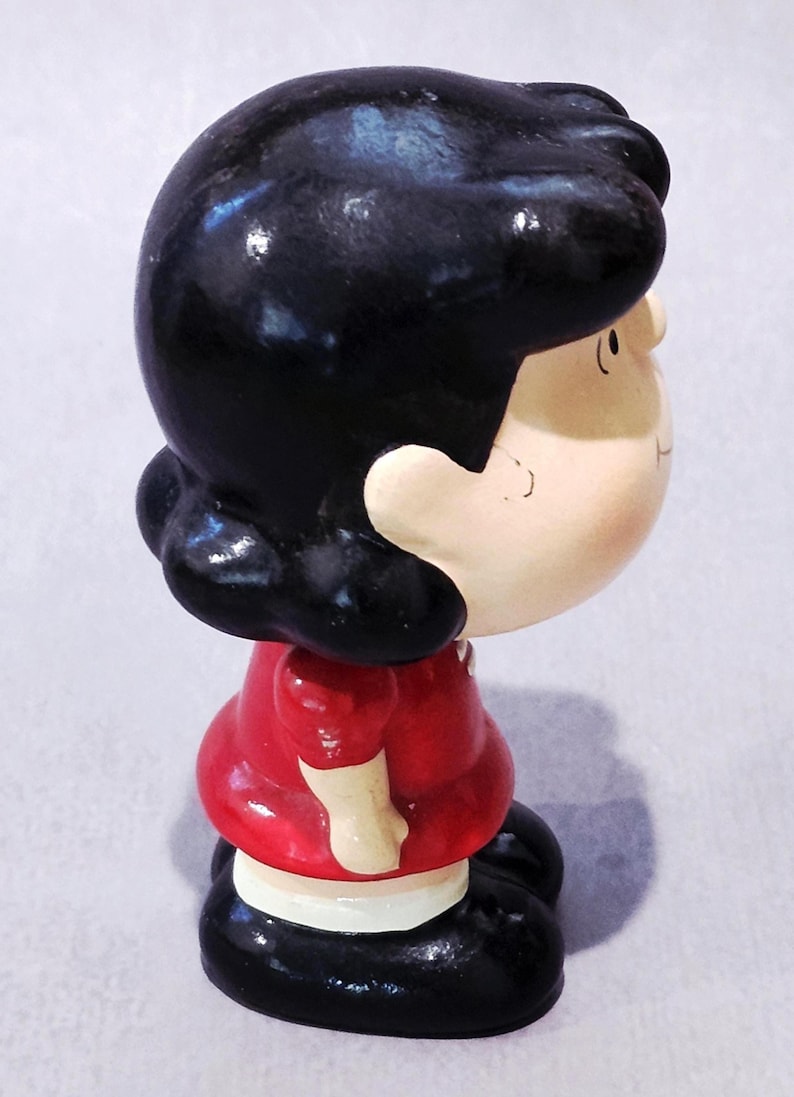 May include: A ceramic figurine of a cartoon character with black hair, a red dress, and black shoes. The figurine is painted with a glossy finish and has a detailed facial expression. The character is looking to the side.