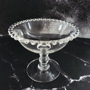 May include: A clear glass dessert bowl with a beaded rim. The bowl sits on a stem with a round base. The glass is transparent, allowing a view of the background. The bowl is likely used for serving desserts or small portions.
