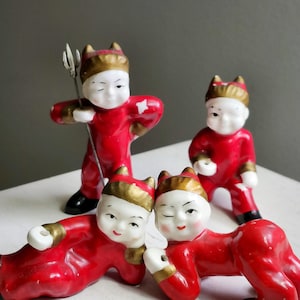 May include: Four small, vintage devil figurines. Each figurine is red with gold accents and a devil horn hat. One figure holds a trident. The figurines are on a white surface.