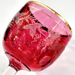 May include: A ruby red wine glass with a gold rim. The glass features a detailed etched design of grape leaves and vines. The stem is clear, and the glass has a rounded bowl shape. The glass is likely made of crystal.