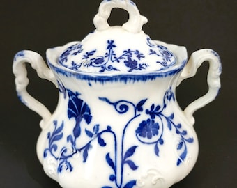 May include: A white porcelain sugar bowl with a lid, featuring intricate blue floral patterns and scrolling vines. The vessel has two ornate, curved handles on opposite sides and a matching lid with a decorative finial handle on top.